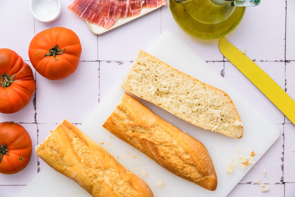 pieces of baguette cut in half on white cutting board with tomatoes and knife around.