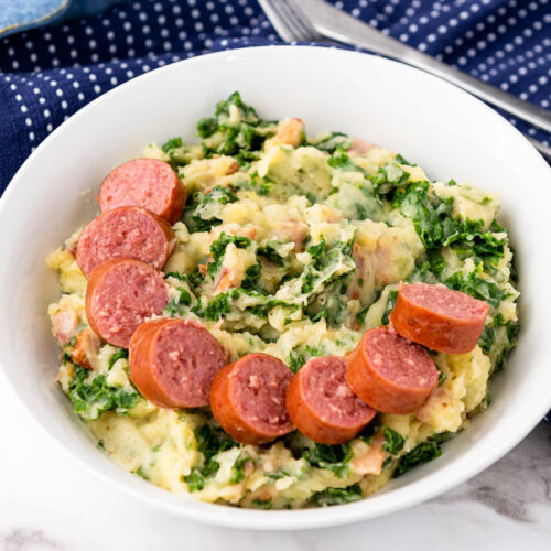 cut up sausage on mashed potatoes with kale in white bowl with blue cloth under and behind.