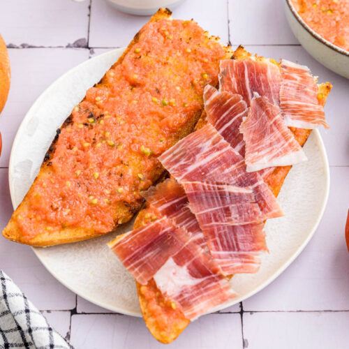 two pieces of baguette sitting on white plate with tomato spread and spices of ham on top.