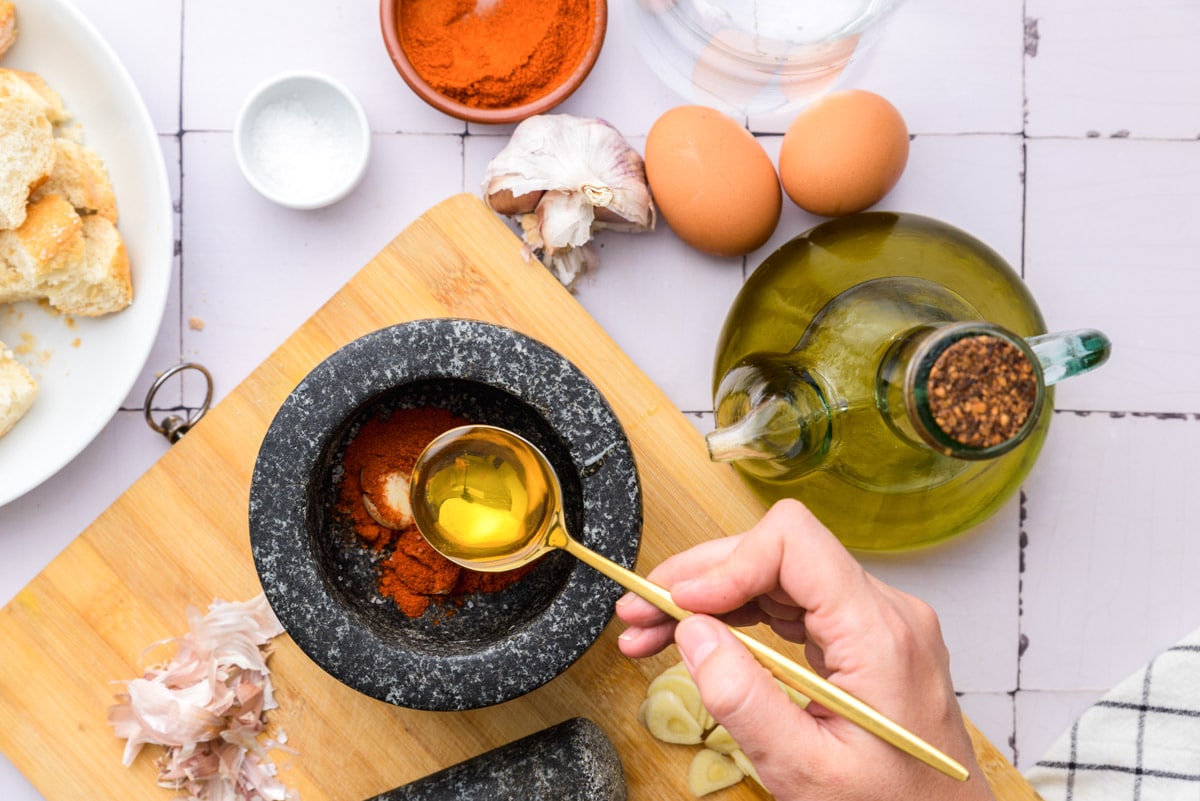 golden spoon with oil held in hand above black mortar and pestle sitting on wooden cutting board with ingredients around.
