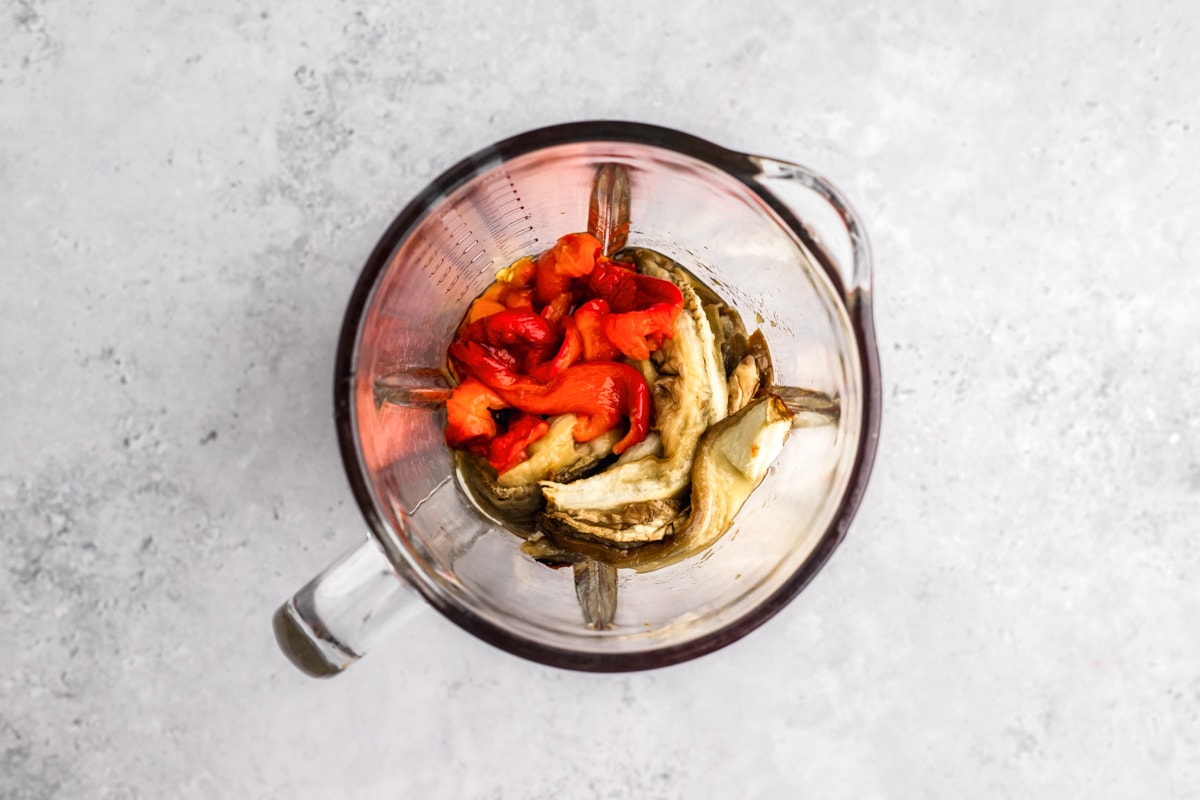 red peppers and eggplant in glass blender on grey counter top.