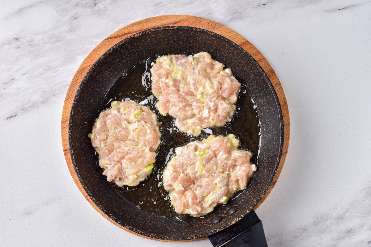 three raw chicken patties in black frying pan with white counter around.