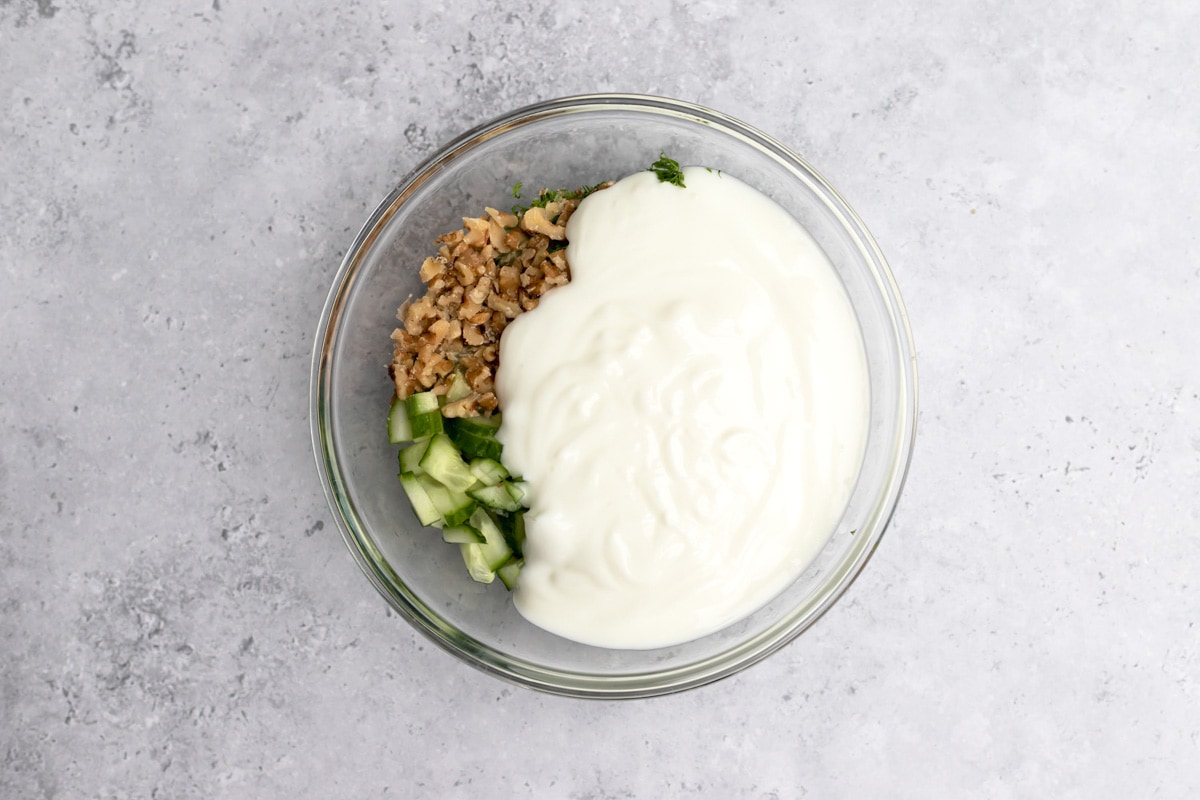 yogurt poured into clear glass mixing bowl with chopped walnuts and cucumber underneath.