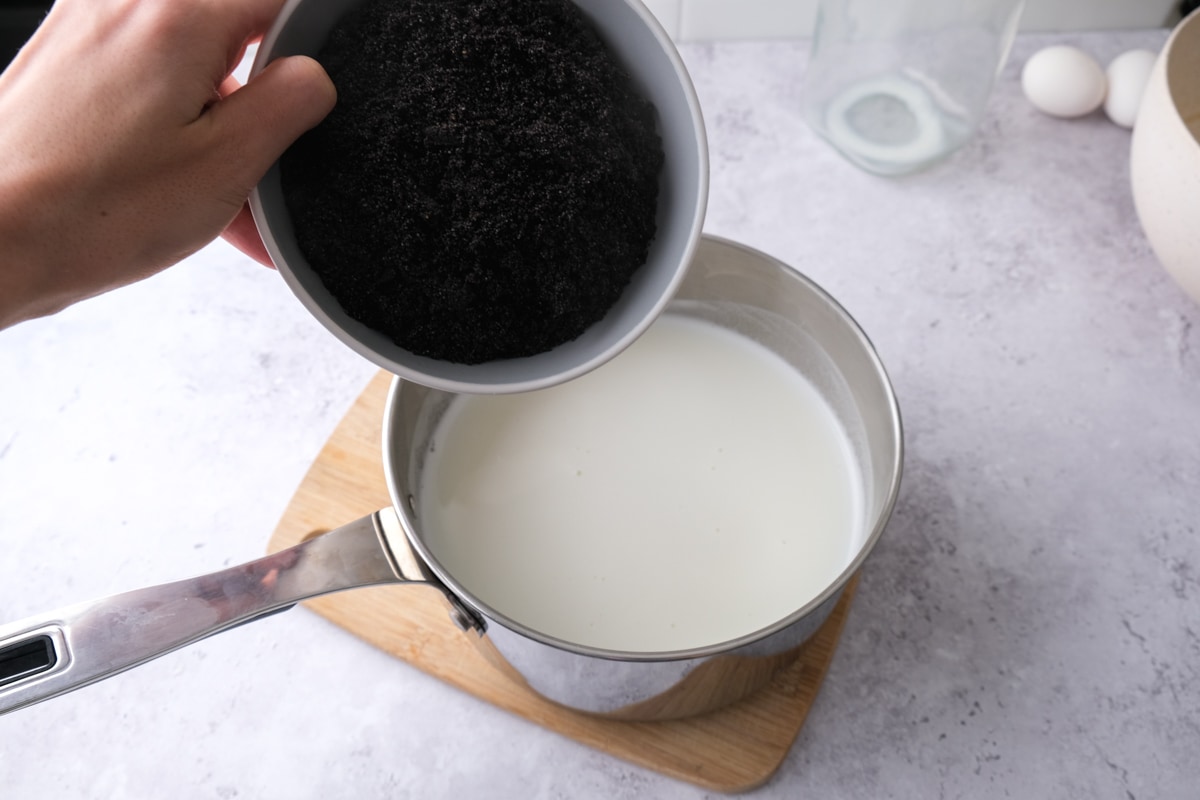 bowl of ground poppy seeds pouring into silver pot of heated milk.
