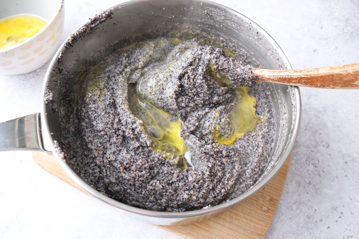 raw egg being mixed into silver pot of poppy seed mixture with wooden spoon.