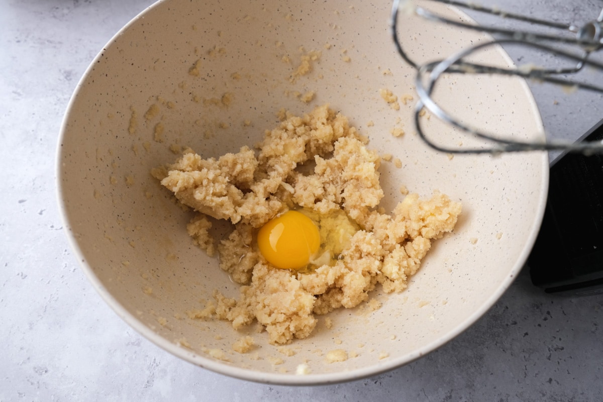 raw egg cracked into bowl of buttery mixture with electric beaters above.