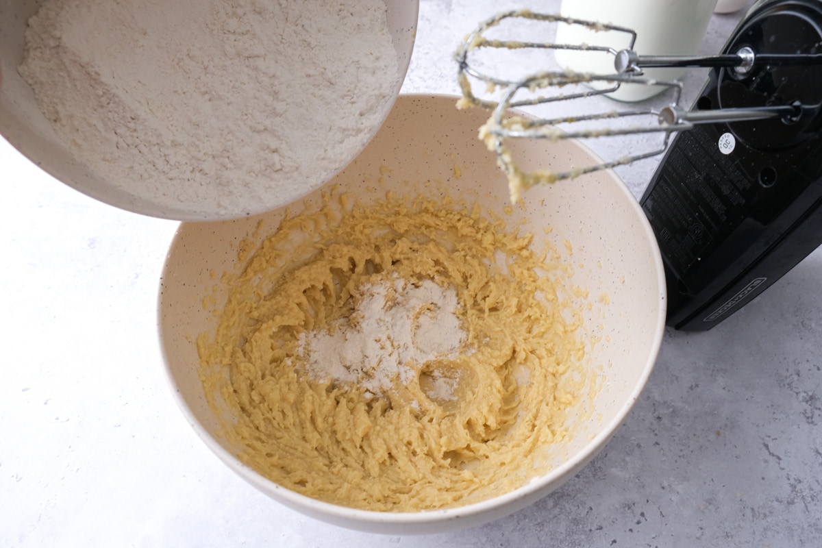 pouring white dry ingredients into bowl of wet buttery batter with electric mixer beside.