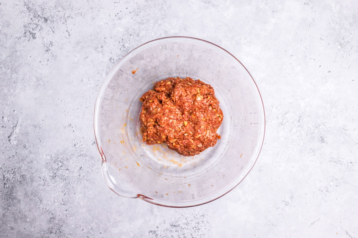 ball of raw meat and spices in clear glass mixing bowl on grey counter.