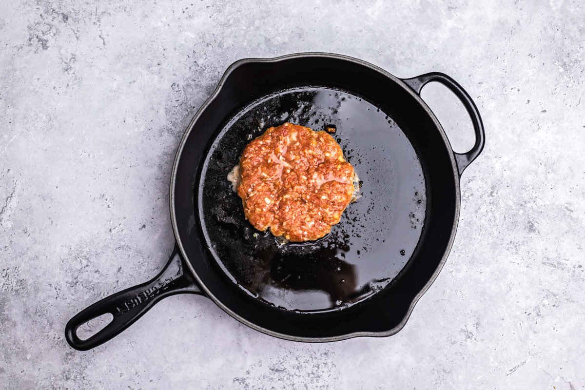round raw meat patty frying in oil in black cast iron pan.