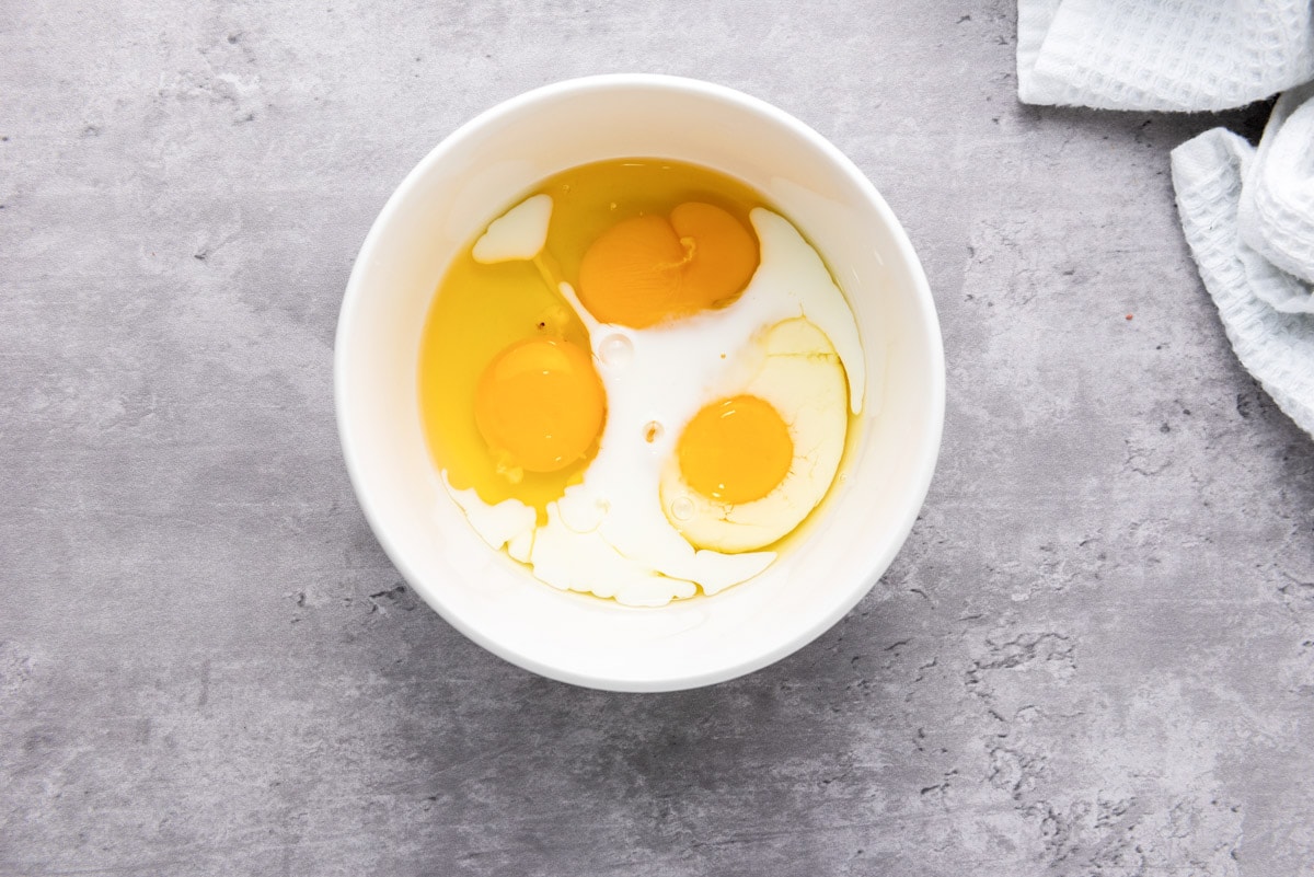 raw eggs cracked into white bowl with milk sitting on grey counter.