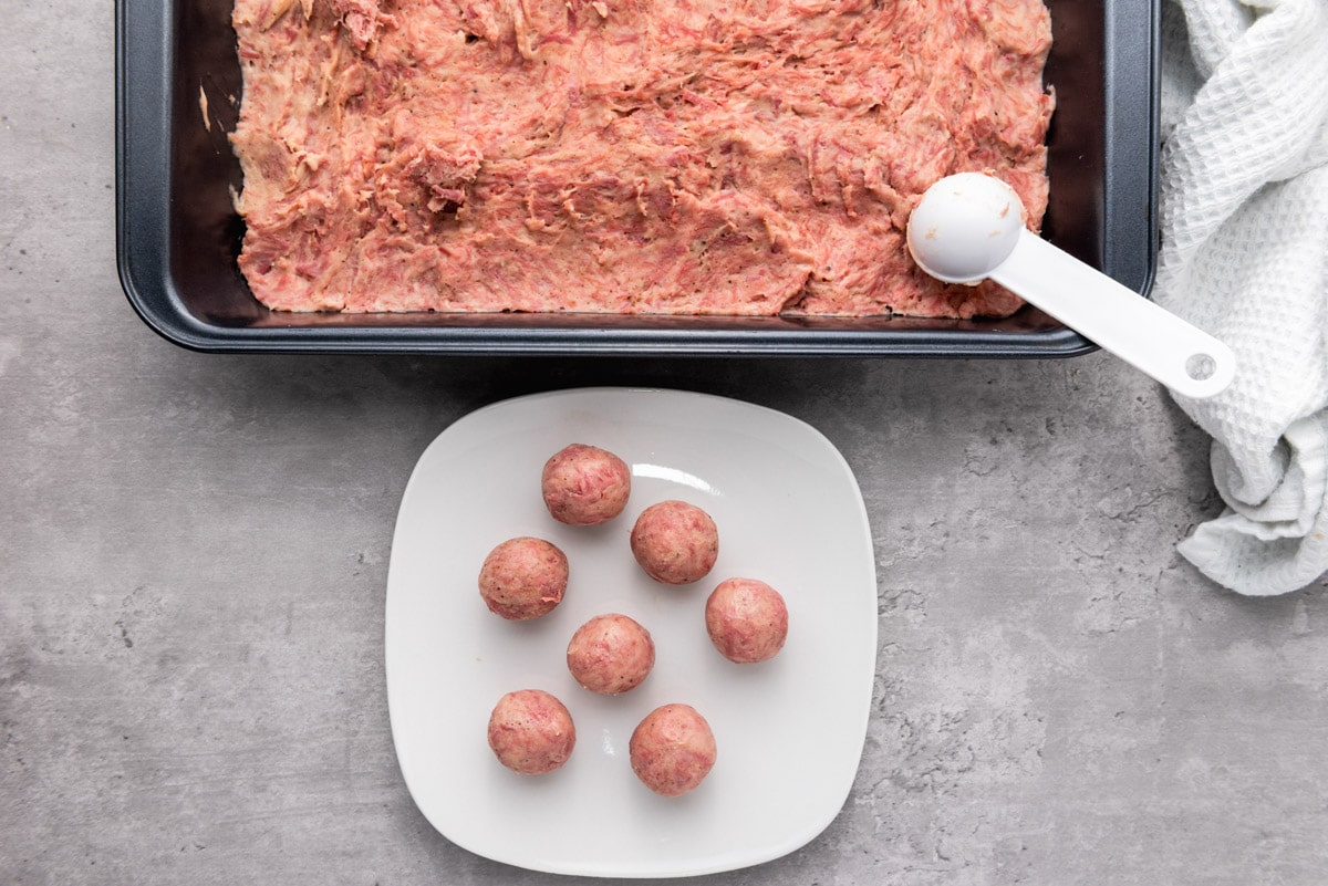 white scoop forming balls of pink meat mixture from large pan of mixture beside.