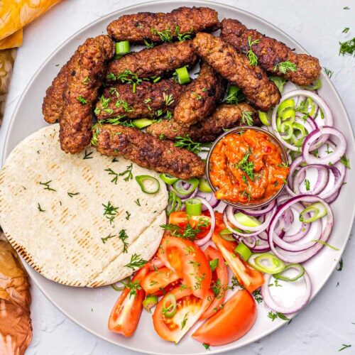 cevapi on plate with ajvar and slices tomatoes and onions.