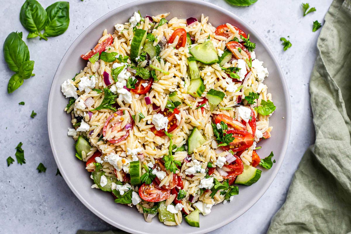 large bowl of colorful orzo salad with basil leaves and green cloth beside.