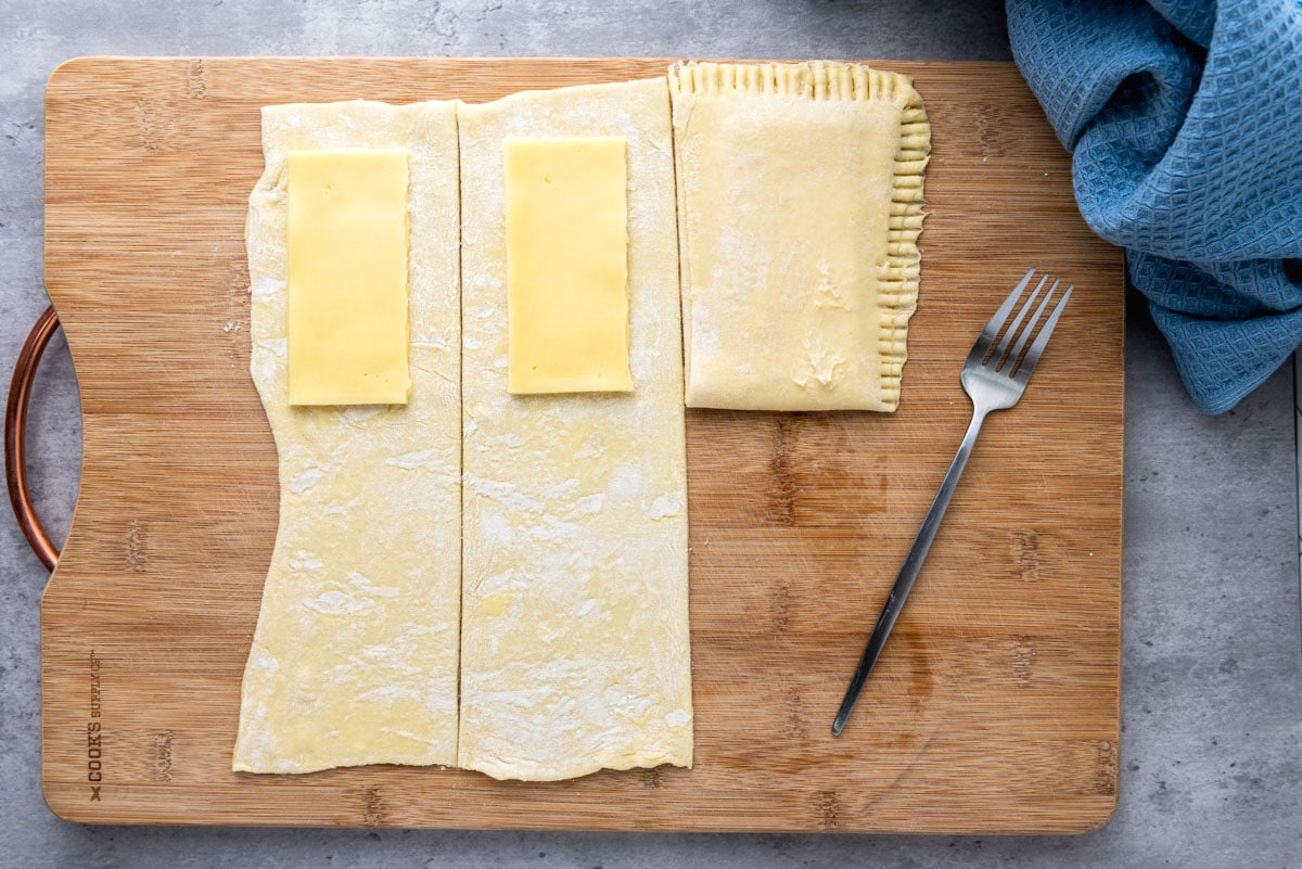 sheets of puff pastry in rows on wooden board with slices of gouda resting on top.