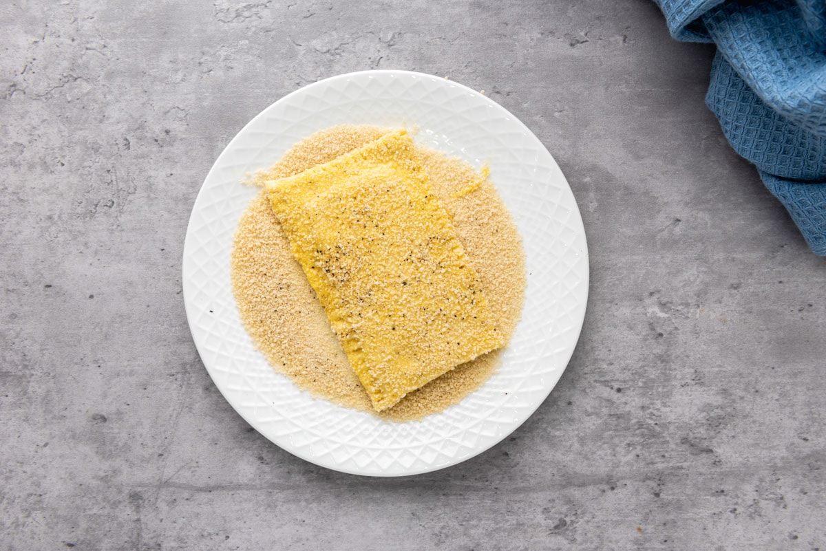 square of puff pastry rolled in breadcrumbs on white plate sitting on grey counter top.