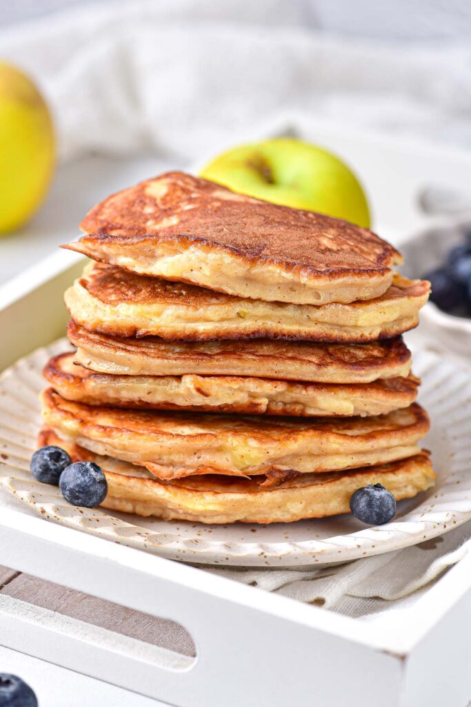 stack of apple pancakes on plate with blueberries in front and green apple behind.
