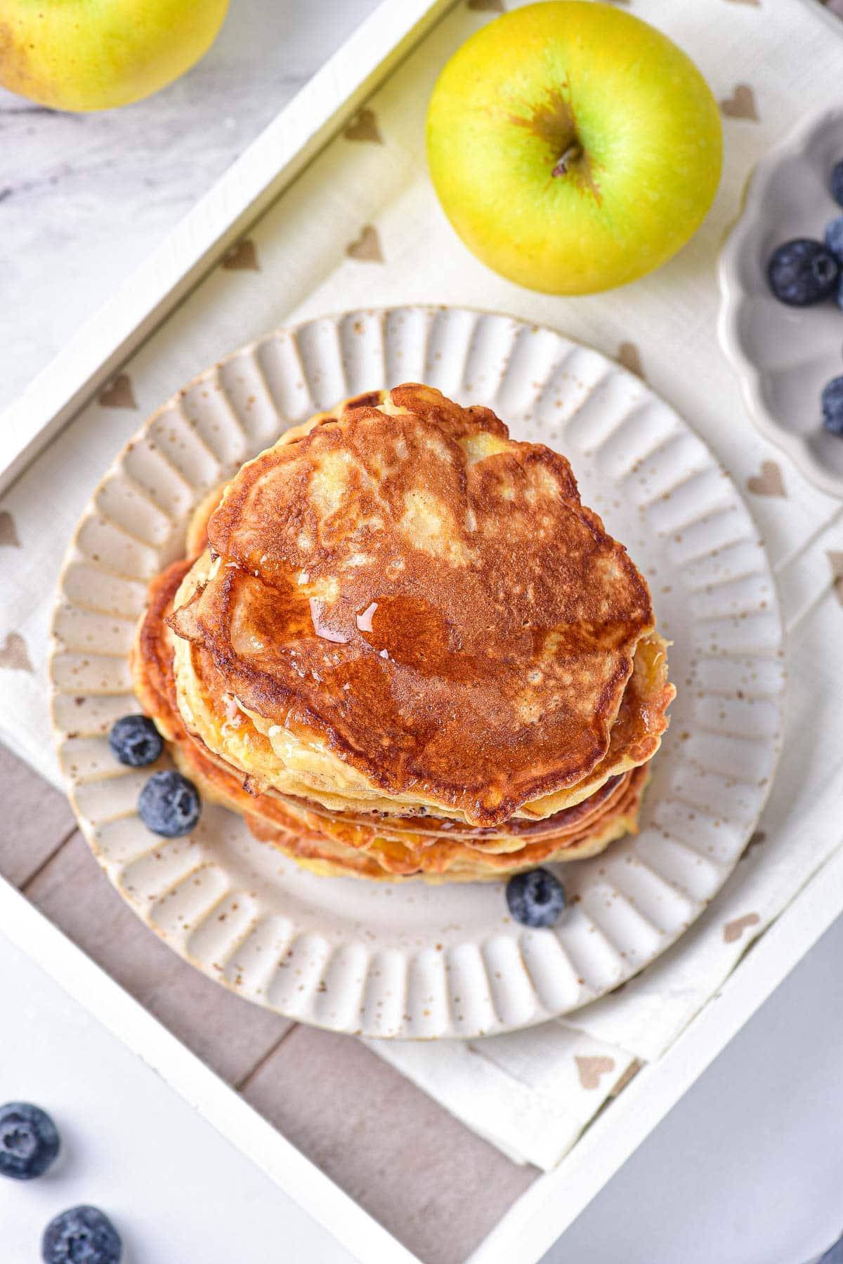 tall stack of golden brown apple pancakes on white plate with berries and apple around.