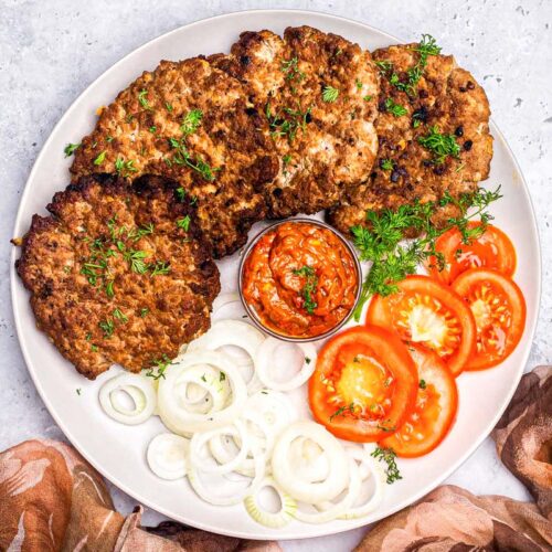 large plate of serbian burger patties with sliced tomatoes and onion.