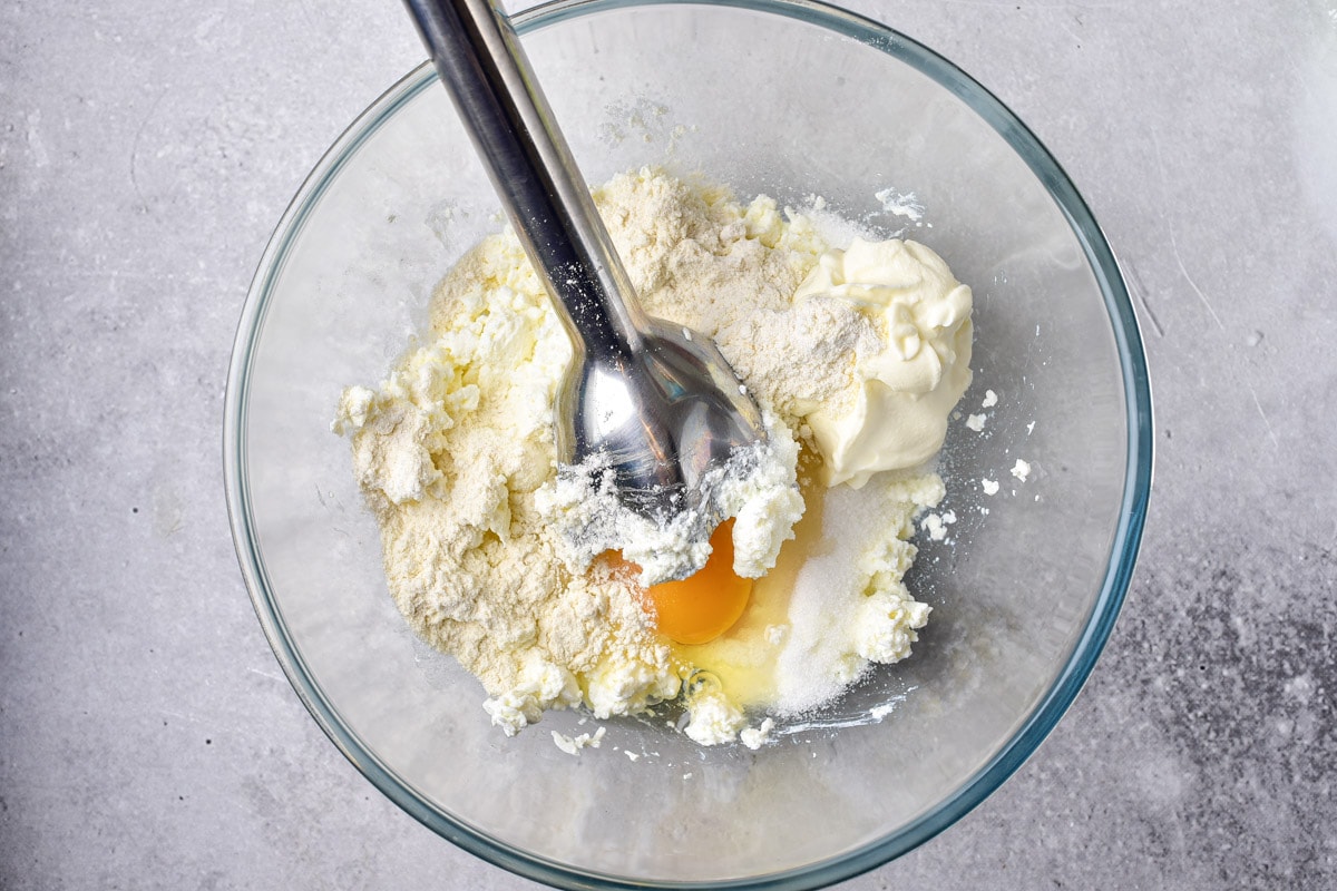 cottage cheese in clear glass bowl with raw egg cracked into being mixed by silver immersion blender.