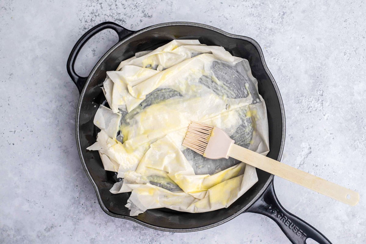 kitchen brush brushing melted butter on phyllo dough in cast iron pan.
