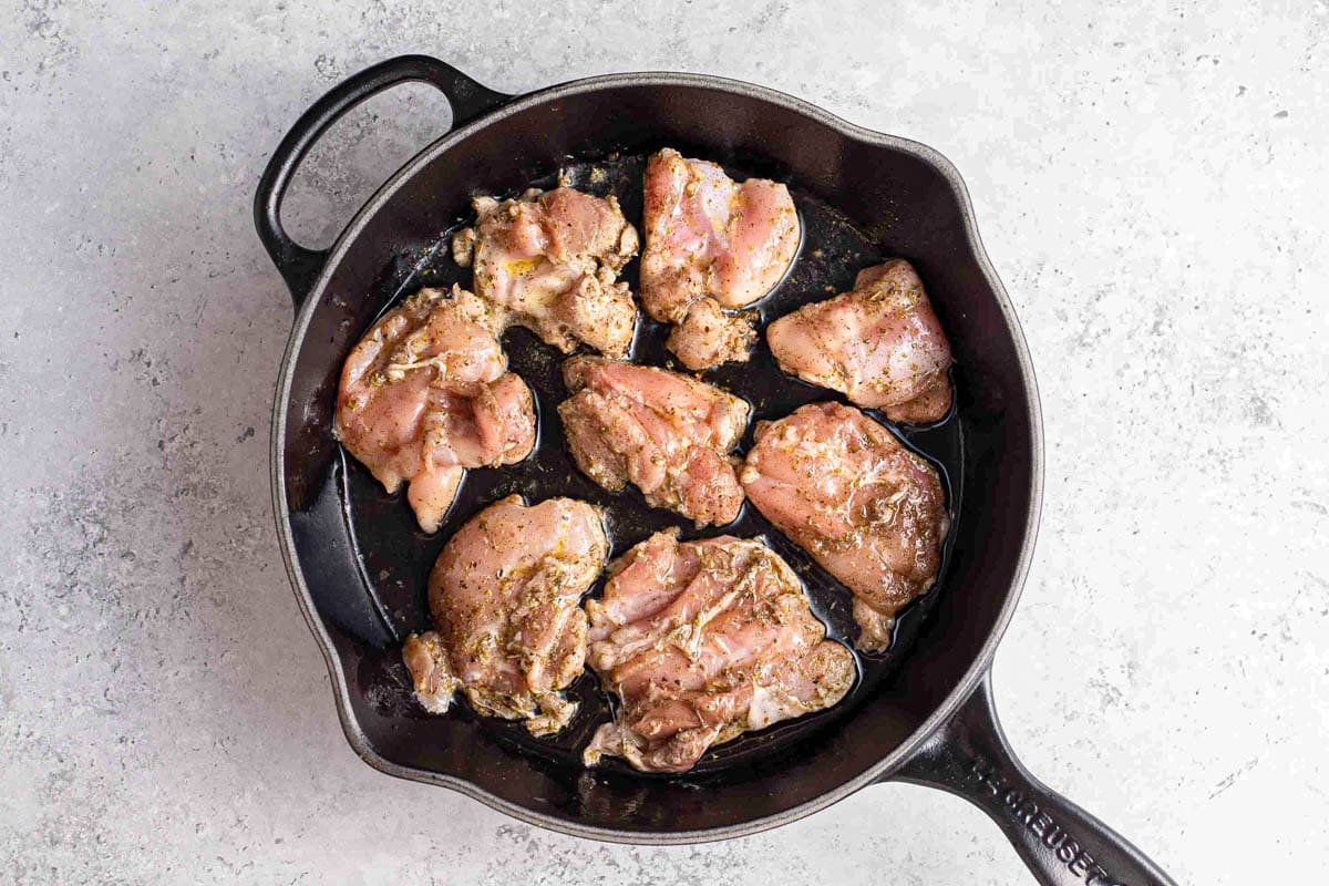 raw chicken thighs cooking in black cast iron pan with grey counter top around.