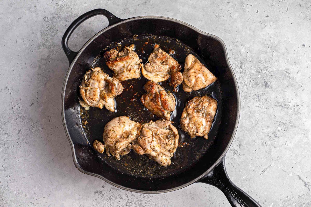 cooked chicken thigs frying in black cast iron pan with grey counter around.