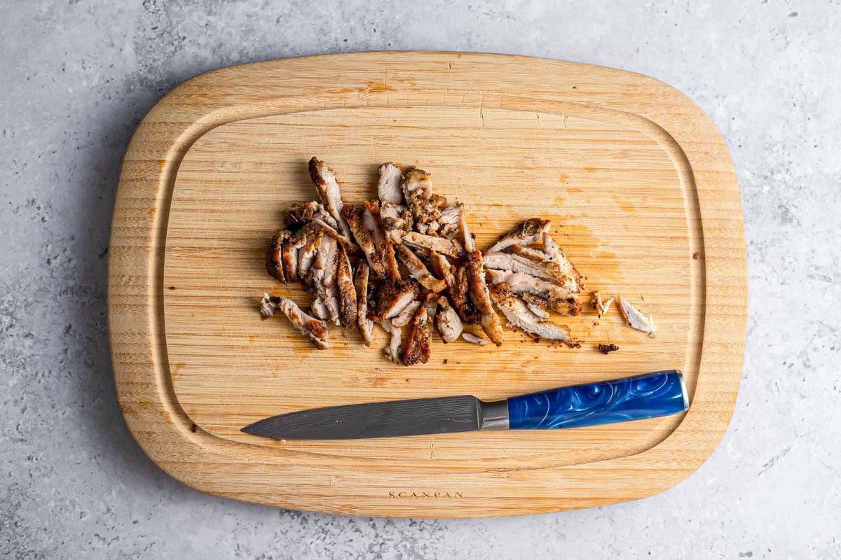 cooked chicken thigh cut into strips on wooden board with knife beside.