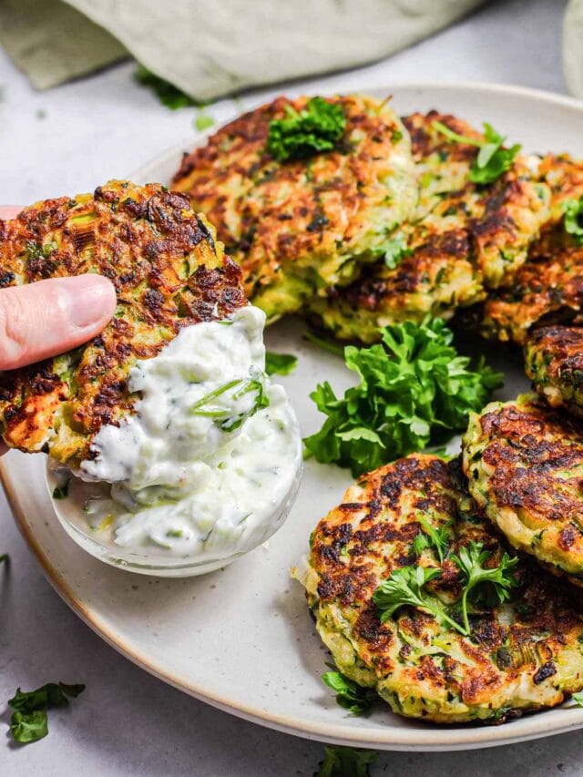 cooked zucchini fritters on plate with hand dipping one in tzatziki sauce.