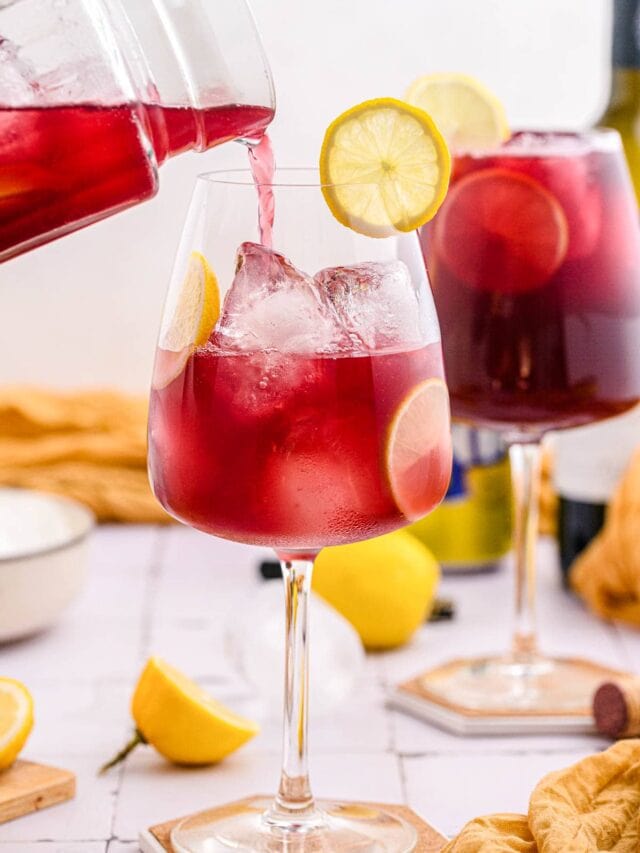 pitcher pouring red wine drink into wine glass with lemon slices around on counter.