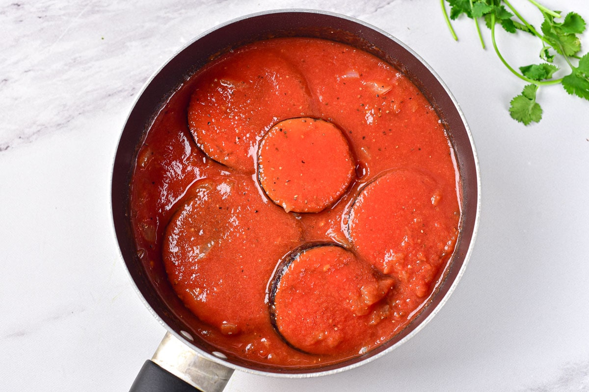 slices of eggplant covered in tomato puree in large black frying pan.