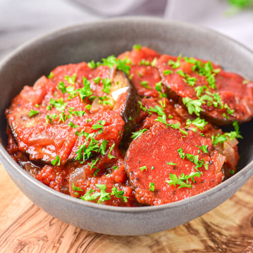 grilled eggplant covered in tomato sauce in grey bowl on wooden board.
