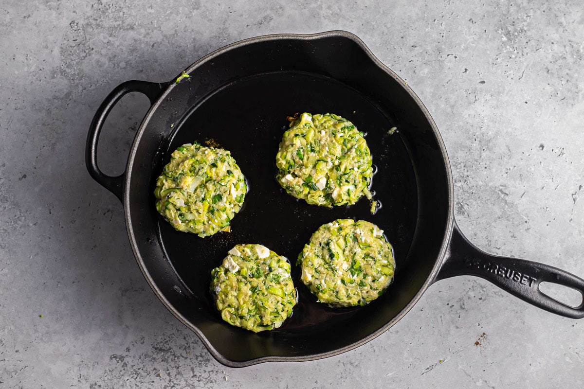 four raw zucchini fritters in black cast iron pan with grey counter around.