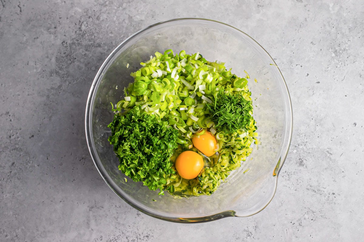 grated zucchini eggs and chopped parsley ingredients in clear glass mixing bowl on counter.