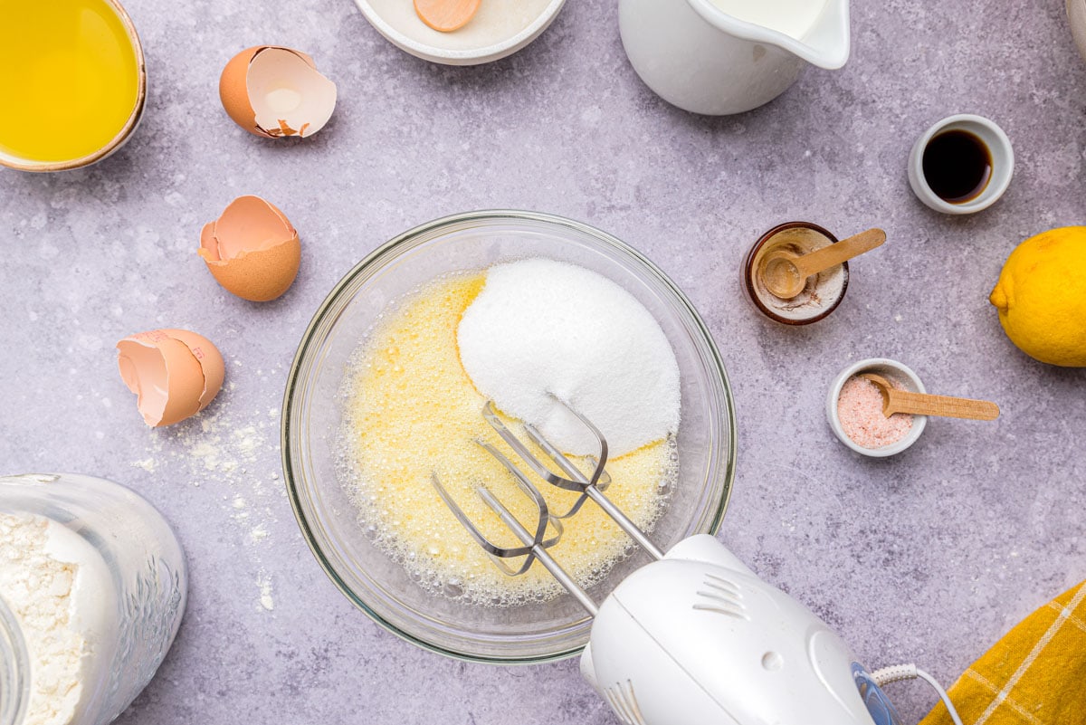 sugar dumped into raw egg mixture in clear glass mixing bowl with electric beaters above.