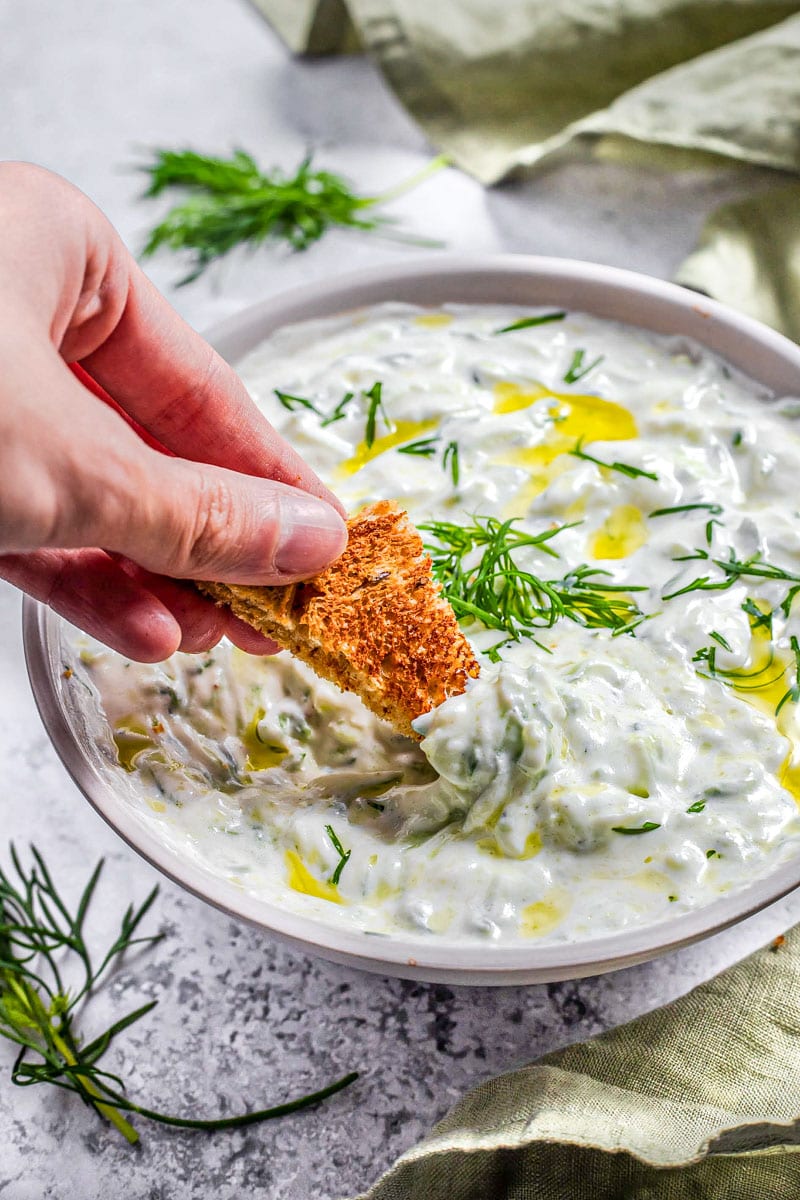 hand dipping piece of bread into large bowl of tzatziki with green cloth beside.