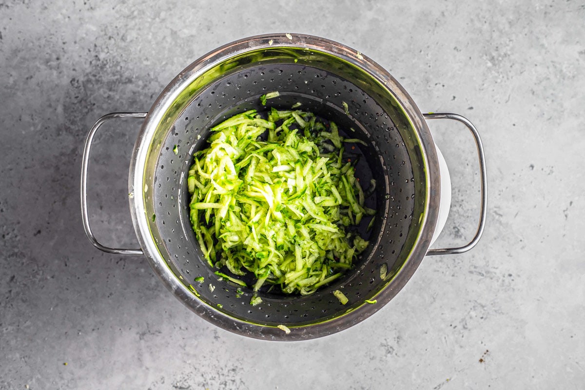 shredded cucumber in silver colander with grey counter top around. 