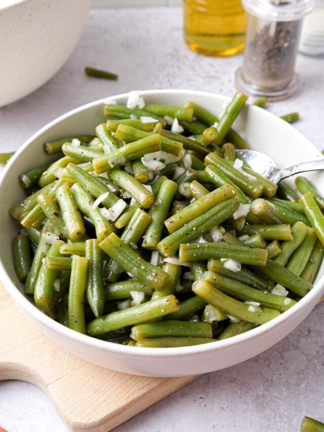 bowl of green bean salad with onion on wooden board on counter.