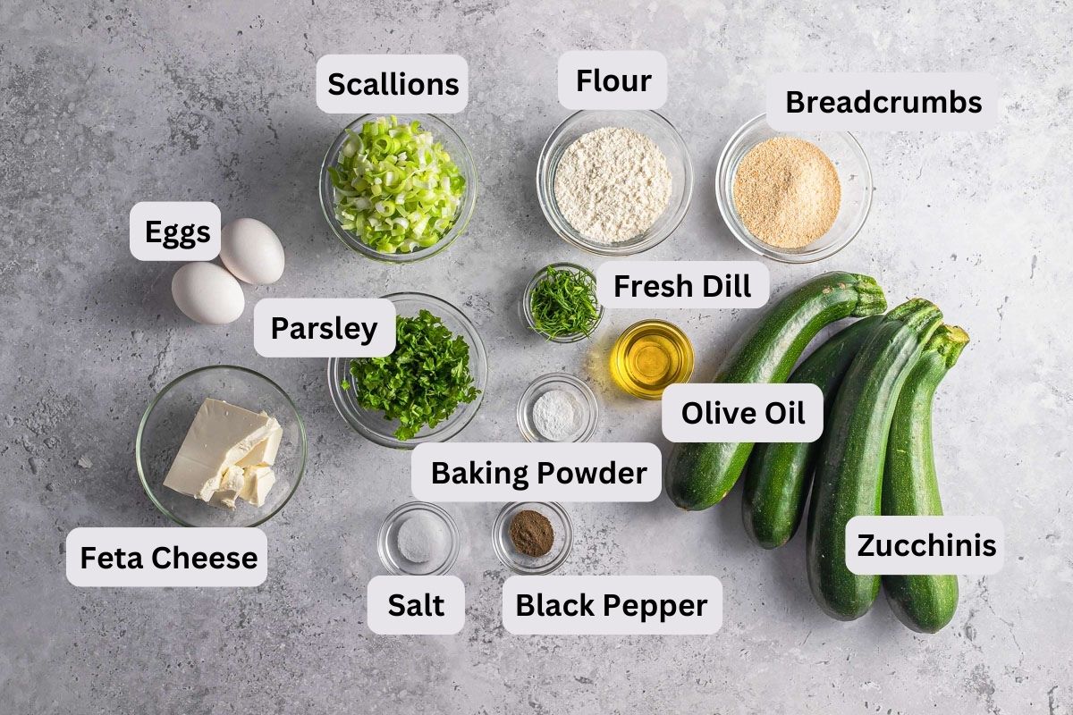 ingredients to make zucchini fritters on grey counter in bowls with labels seen from above.