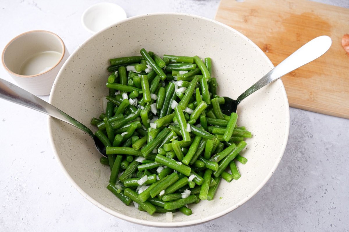green beans in white mixing bowl being tossed by silver spoons in dressing.