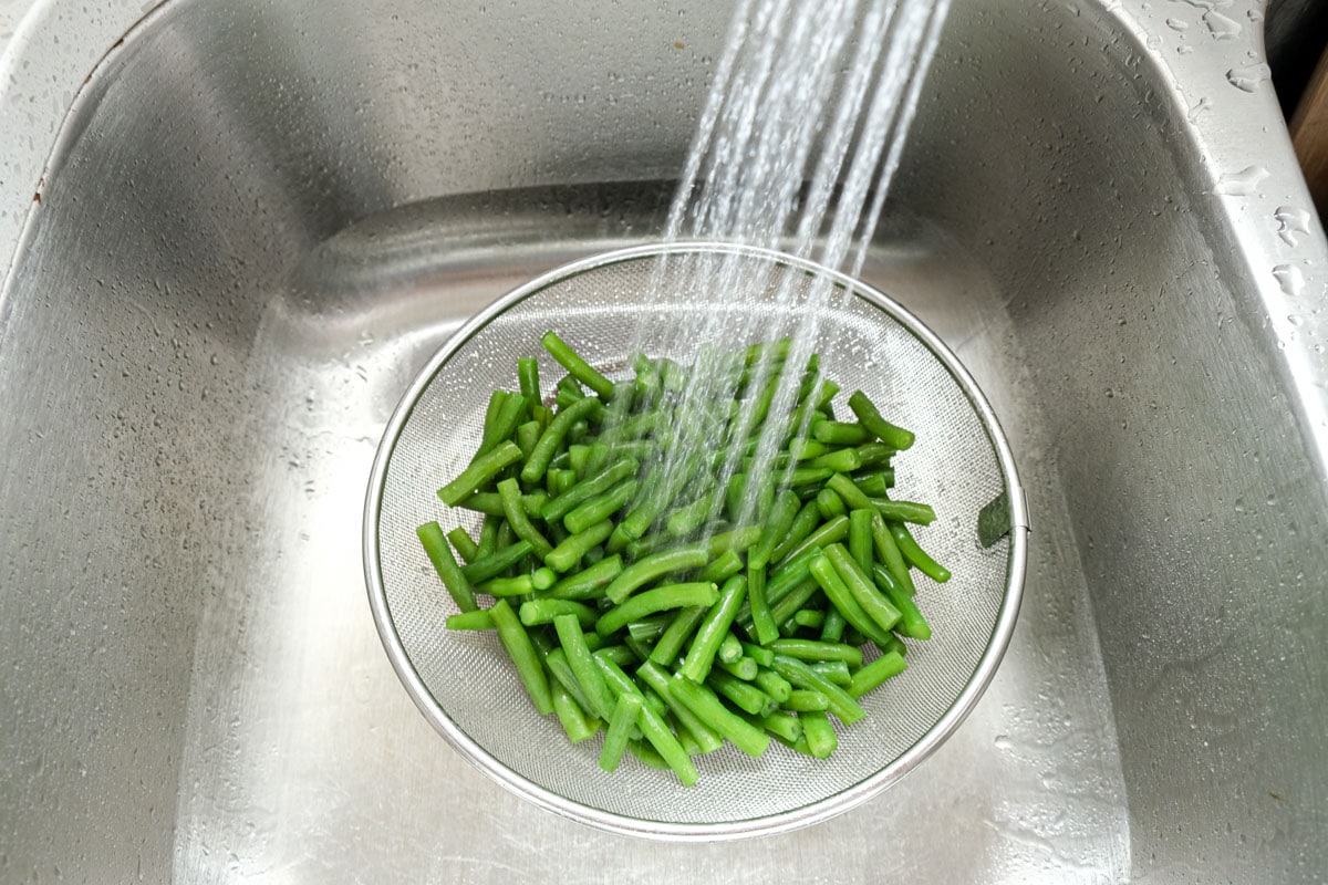 green beans in silver colander being rinsed by running water in sink.