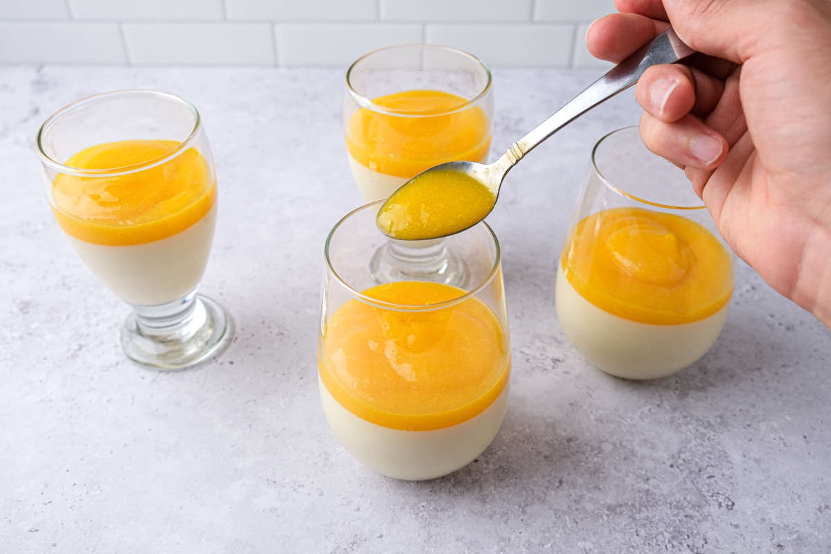 spoon putting mango coulis on top of firmed panna cotta in glasses.