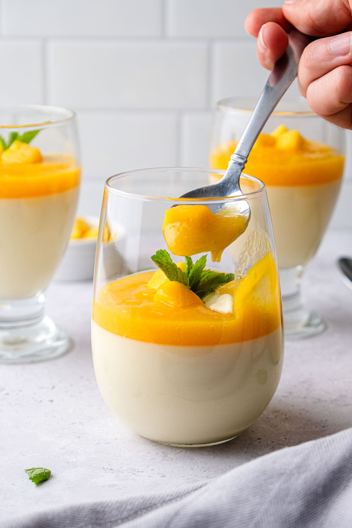 spoon scooping mango panna cotta out of clear glass sitting in grey counter.