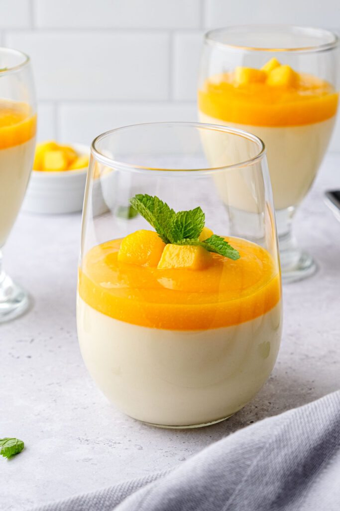 clear glass of mango panna cotta with mint leaves and other desserts behind.