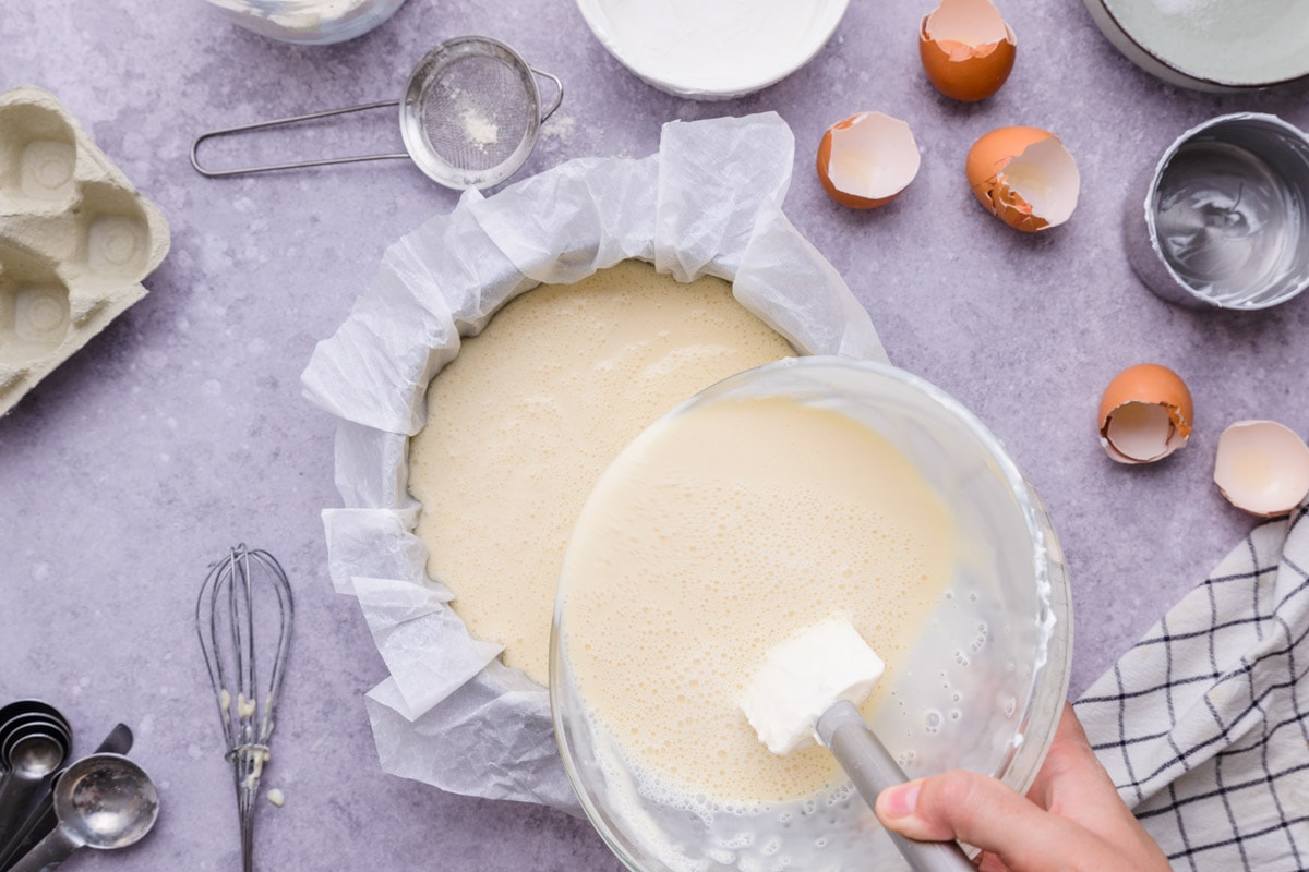 white spatula pouring batter from mixing bowl into cake form lined with parchment paper.