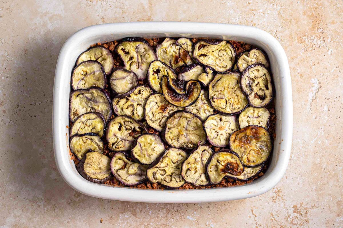 layer of grilled eggplant on meat in white rectangle baking dish.