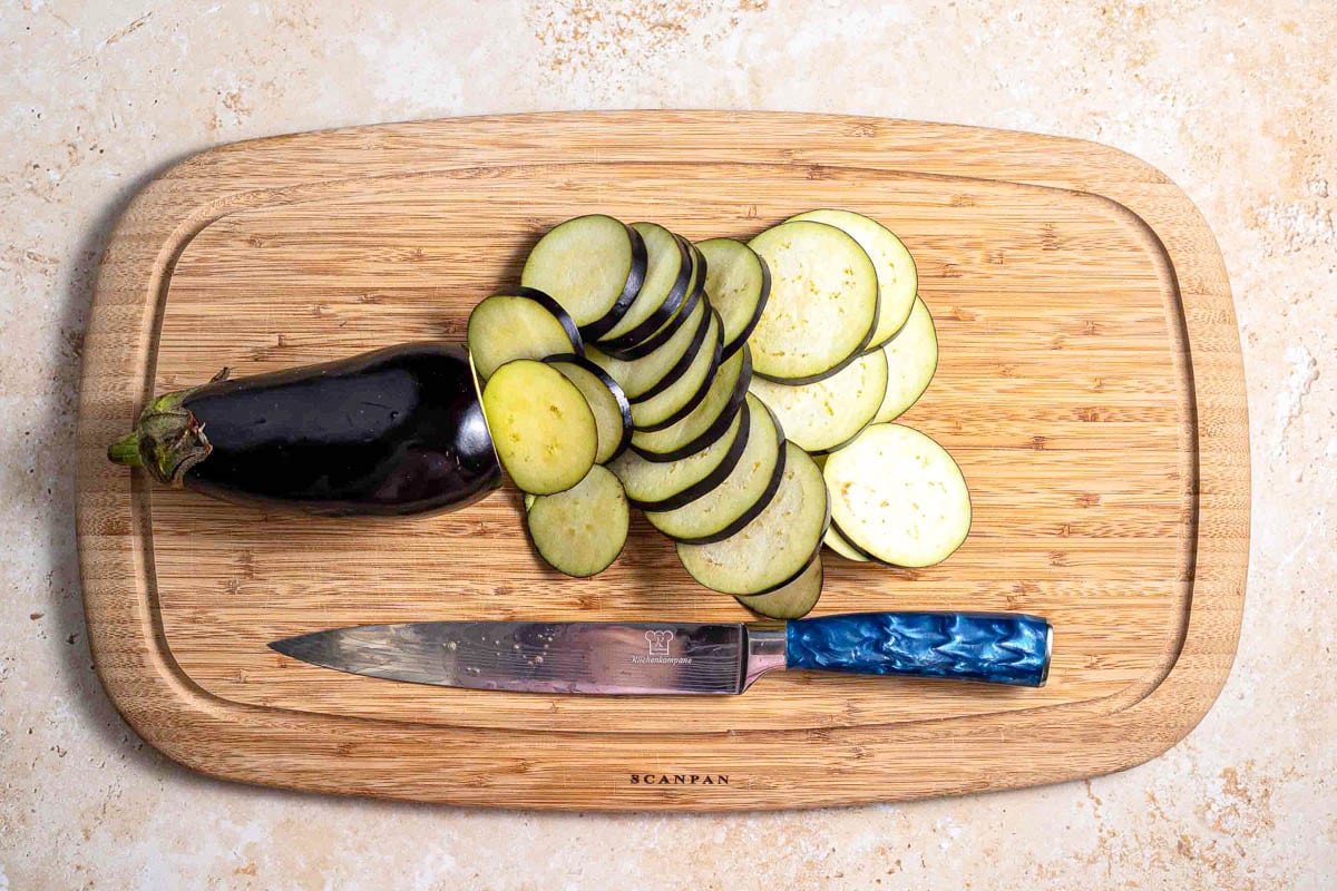 knife on wooden cutting board beside thinly sliced eggplant.