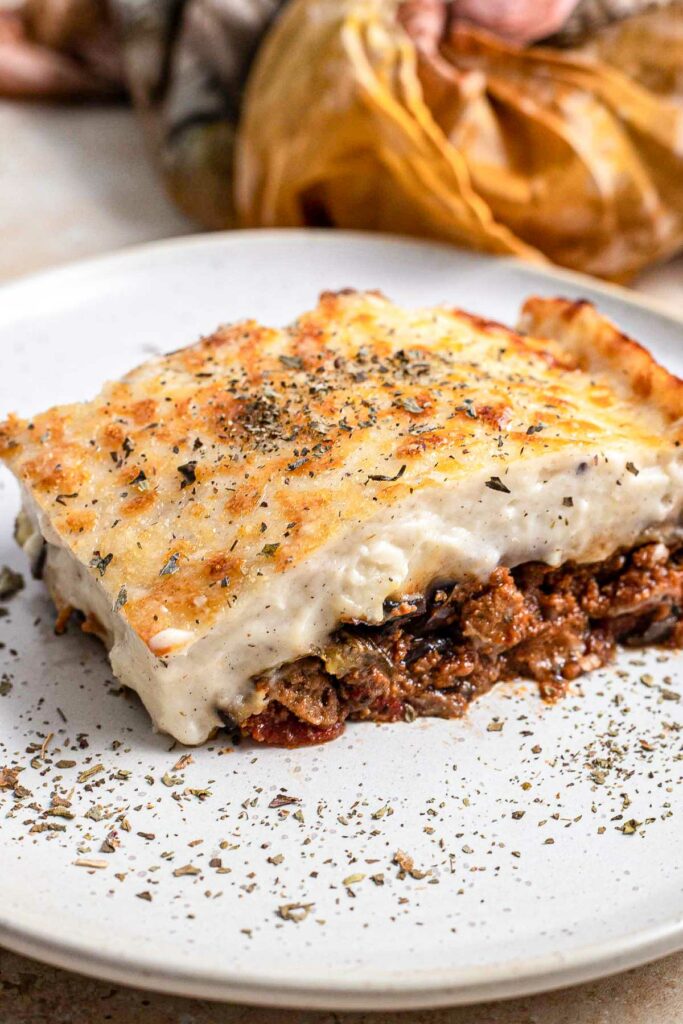 slice of moussaka on white plate with spices around and cloth behind.