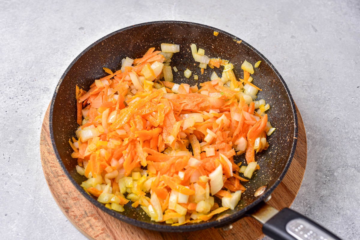 shredded carrots and chopped onions in frying pan sitting on wooden pot holder.