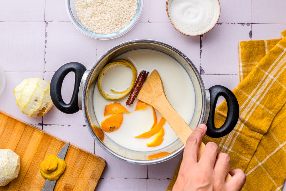hand holding wooden spoon stirring milk and citrus peels in silver pot on counter.