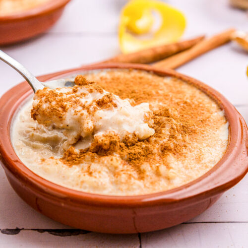 red dish of spanish rice pudding covered in cinnamon with silver spoon removing some.